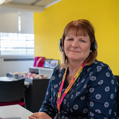 A professional woman sits at a desk in an office with a bright yellow wall behind her. She's wearing a dark patterned top and is positioned at a workstation with a computer monitor.