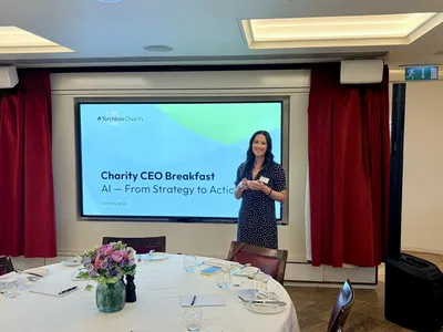 Lisa stands smiling beside a large presentation screen displaying “Torchbox Charity – Charity CEO Breakfast: AI — From Strategy to Action” dated 13th May 2026. She is wearing a dark polka-dot dress and holding a cup and saucer. In front of her is a round breakfast table set with flowers, notebooks, glasses, and place settings, while red curtains frame the presentation area.