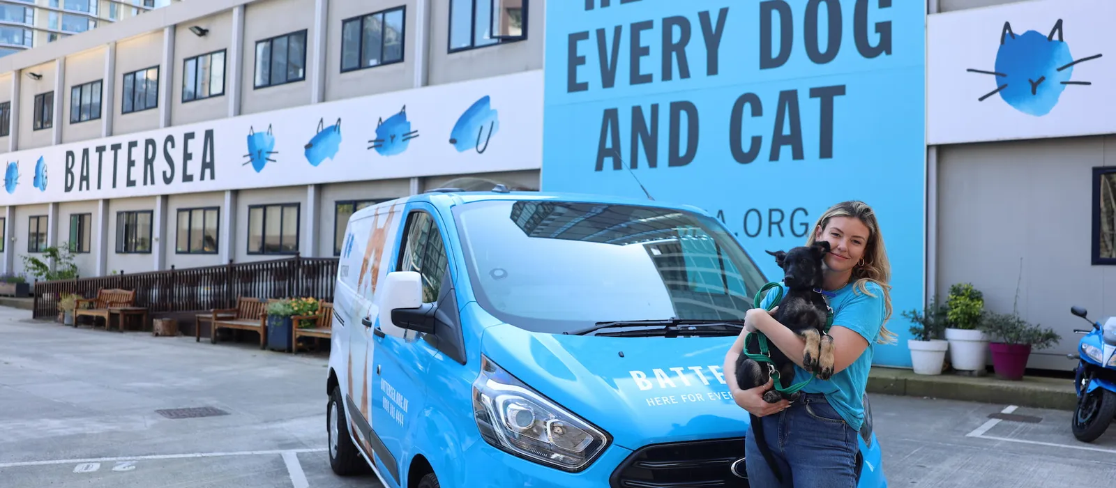 Battersea staff member standing outside a Battersea building, holding a small black dog beside a blue van. Large sign behind reads “Here for every dog and cat”.