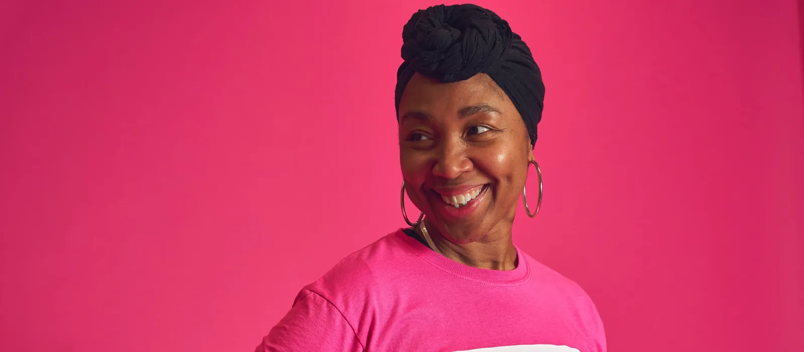 A smiling person wearing a bright pink Breast Cancer Now t-shirt and black headwrap stands against a pink background, looking to one side.