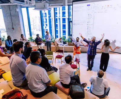 A group of people sit and stand in a bright, modern meeting space with tiered seating, watching three presenters at the front of the room. Large windows overlook nearby buildings, and a projected computer screen is displayed on the wall behind the presenters.