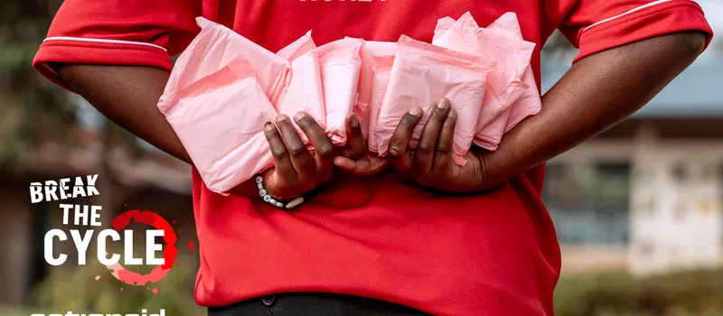 A close up shot of a person in a red ActionAid shirt holds bundles of pink sanitary pads. Text reads “Break the Cycle,” alongside the ActionAid logo.