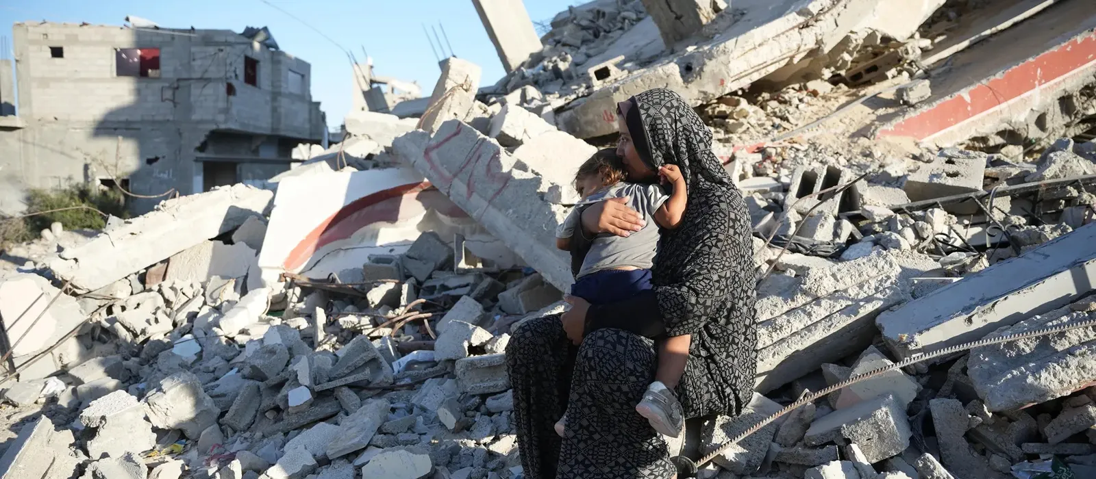 A woman sits holding a small child among the rubble of collapsed buildings and broken concrete, looking out across the destruction in a heavily damaged urban area.