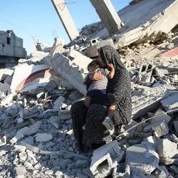 A woman sits holding a small child among the rubble of collapsed buildings and broken concrete, looking out across the destruction in a heavily damaged urban area.