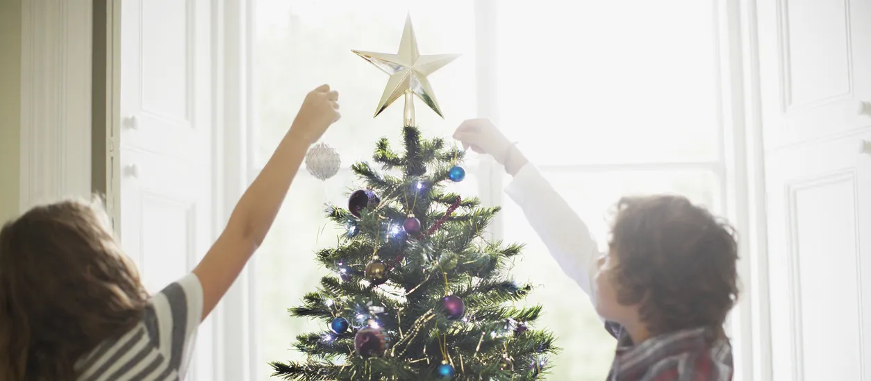 Two children decorating a Christmas tree with colourful baubles and fairy lights, topped with a gold star.
