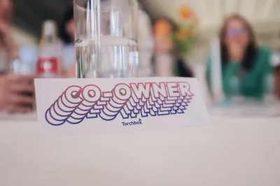 A table name card displaying the words “Co-Owner” in colourful layered typography, photographed on a meeting table with Torchbox colleagues blurred in the background.