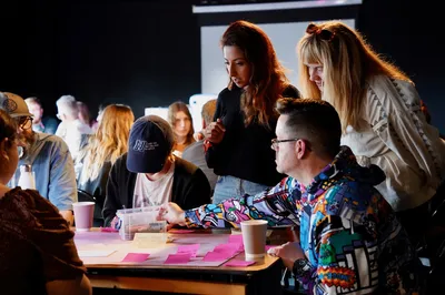 A group of people sit and stand around a table covered with pink sticky notes, paper, and coffee cups during a workshop. One person reaches into a box of supplies while others lean in to discuss ideas. The room is softly lit, and several other groups are visible in the background, suggesting a collaborative, creative session.