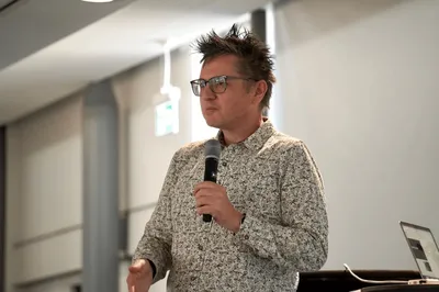 Nick wearing glasses and a patterned shirt speaks into a microphone in a bright, modern room. A laptop sits open on a nearby table.