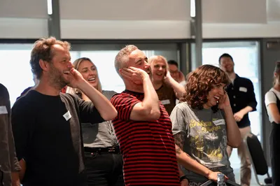 Ben, Miles, and Cassie laugh together during a lively laughter yoga session, surrounded by colleagues who are all smiling and joining in. The room feels bright, relaxed, and full of energy.