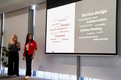 Lily and Lisa stand on stage in front of a large presentation screen displaying words related to both the charity and public sectors.