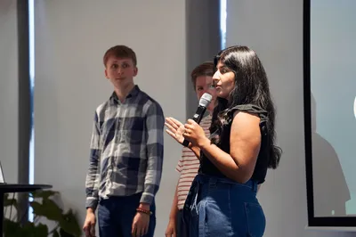 Jess speaks into a microphone while presenting on stage, with James and Holly standing beside her and listening. The three colleagues share the stage in a bright, relaxed setting filled with natural light.