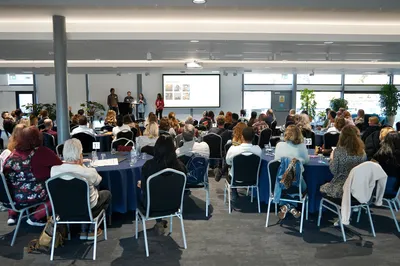 A wide shot of the Torchbox team gathered in a bright, open room. Colleagues sit at round tables listening to a group presenting at the front, where a slide is displayed on a large screen. The atmosphere feels relaxed and collaborative.