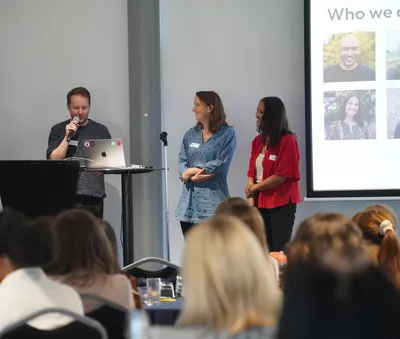 Jonny, Helen and Lisa stand at the front of a room speaking to Torchbox co-owners during a company day presentation. Jonny is speaking into a microphone while Helen and Lisa stand beside him.