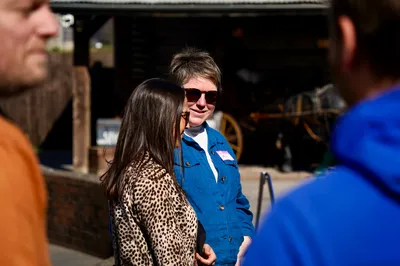 Holly and Lisa talk outdoors on a sunny day, Lisa wears a leopard print jacket, Holly wears a blue denim jacket and sunglasses, with a rustic wooden building in the background.