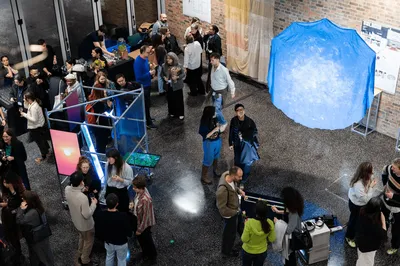 A busy indoor exhibition, with groups of people gathered around artworks and installations. Visitors are chatting, holding drinks, and exploring digital screens and sculptural pieces displayed throughout the space. The scene is lively and informal, set against brick walls and large windows.