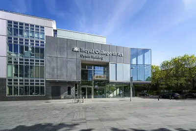 The exterior of the Royal College of Art’s Dyson Building, shown on a bright day with a clear blue sky. The modern structure features large glass panels and metal cladding, with the building name displayed prominently above the entrance. A quiet, open courtyard sits in front, with a bicycle parked near the doorway.