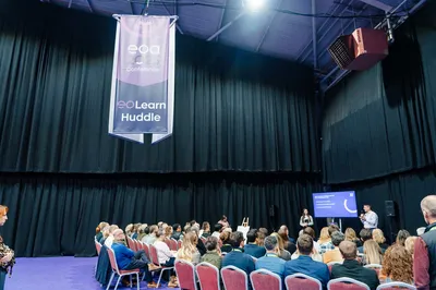 A breakout session at the EOA 2025 Conference, with attendees seated in rows facing speakers and a screen, beneath a hanging “EO Learn Huddle” banner.