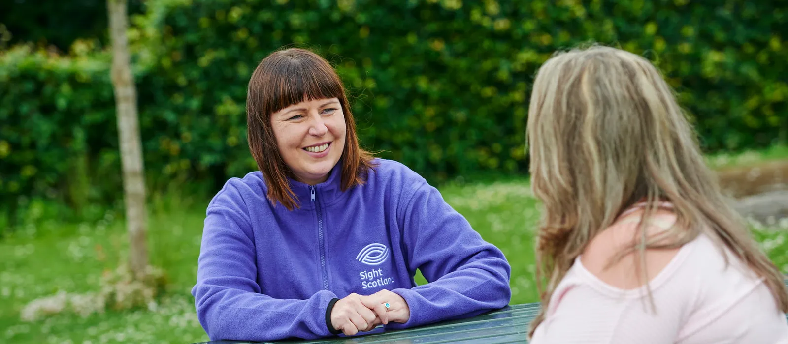 A support worker wearing a purple Sight Scotland fleece sits at an outdoor picnic bench, smiling and talking with another woman. They are in a green park setting with grass and trees in the background.
