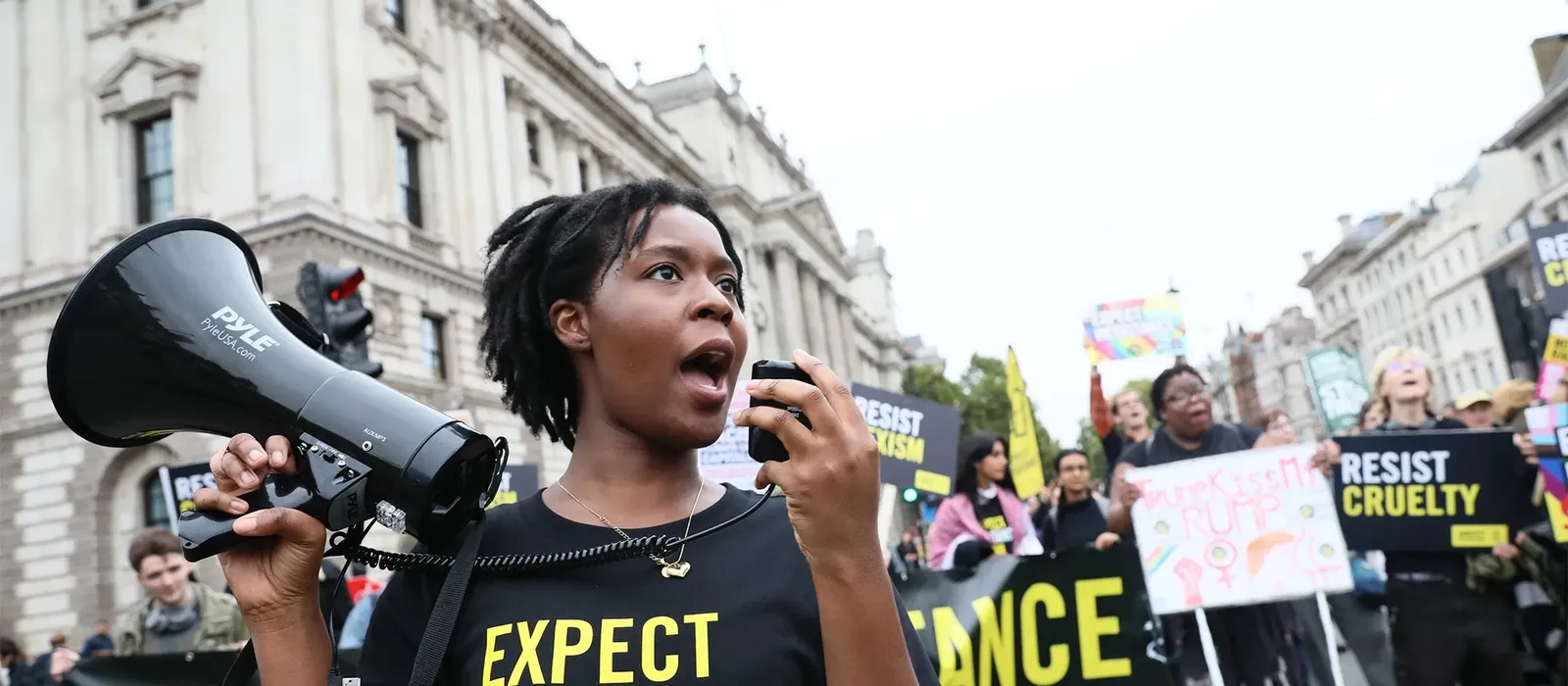 Photograph of a protest scene. A woman speaks into a handheld microphone while holding a megaphone. She is wearing a black T-shirt with the words “Expect Resistance.” Behind her, a crowd hold up signs with the same wording.