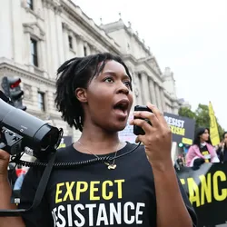 Photograph of a protest scene. A woman speaks into a handheld microphone while holding a megaphone. She is wearing a black T-shirt with the words “Expect Resistance.” Behind her, a crowd hold up signs with the same wording.
