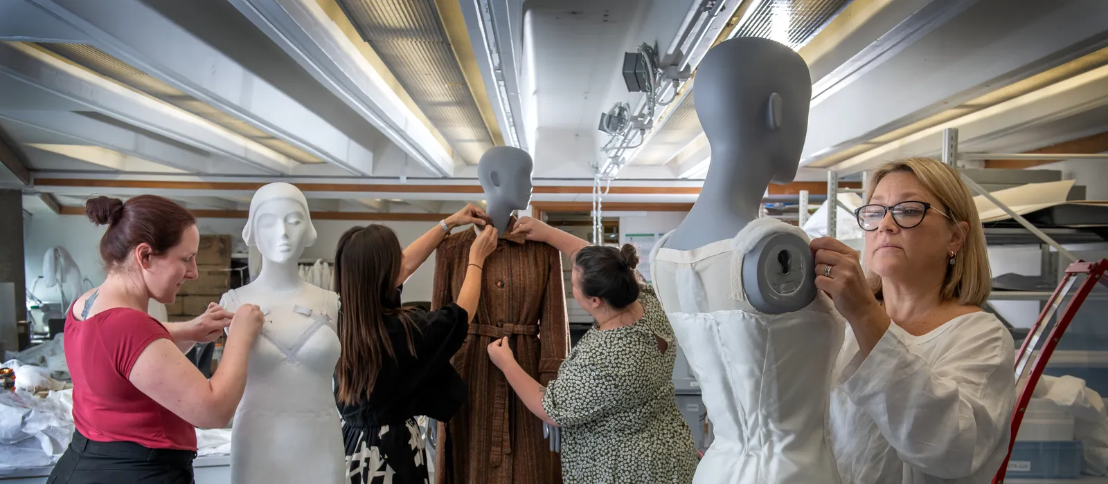 Four women actively engage in conserving and dressing mannequins with historical-looking garments in a well-lit workshop. From left to right, a woman in a red top pins a dress on a mannequin, another two women adjust a long brown dress on a mannequin, and