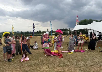 A lively outdoor gathering of Torchboxers. Alex and Naomi are dressed in colourful, playful costumes on a grassy field under a cloudy sky. Bunting, flags and a big white marquee tent set a fun, festival-like atmosphere. Children and adults mingle casually, creating a warm, joyful community vibe.