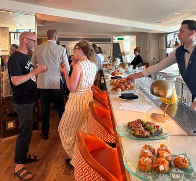 Guests chat and network around a breakfast buffet before the event begins. A long counter is laid out with pastries, fresh fruit, juices and savoury breakfast canapes, while a server offers food to attendees gathered in conversation in a bright, informal reception space.