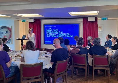 Dr Oonagh Murphy speaks to attendees. She stands beside a large presentation screen displaying the title "What does responsible AI actually mean for cultural institutions?"