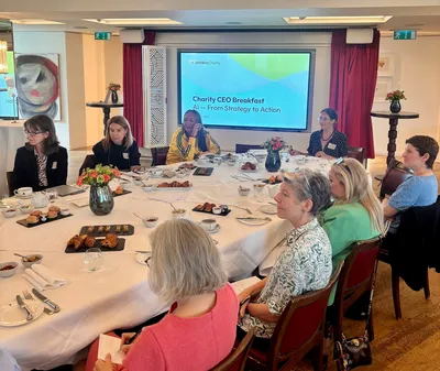Guests sit around a large round table, with pastries, coffee and flowers laid out in front of them. A presentation screen behind the group displays the event title, “Charity CEO Breakfast: AI – From Strategy to Action.” Attendees are listening and taking notes in a bright meeting room.