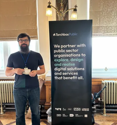 Dan standing beside a Torchbox Public banner at UK Gov Camp, holding a cup of coffee inside Birmingham City Council House.