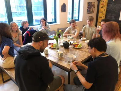 A group of Torchbox co-owners sit around a long wooden table in the Bristol office, chatting over breakfast. Fresh fruit, mugs and a teapot sit in the middle of the table, and people are smiling and talking in small groups.