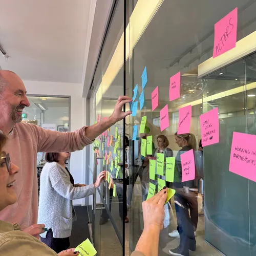 Participants stand along a glass wall, smiling and placing sticky notes as they work together.