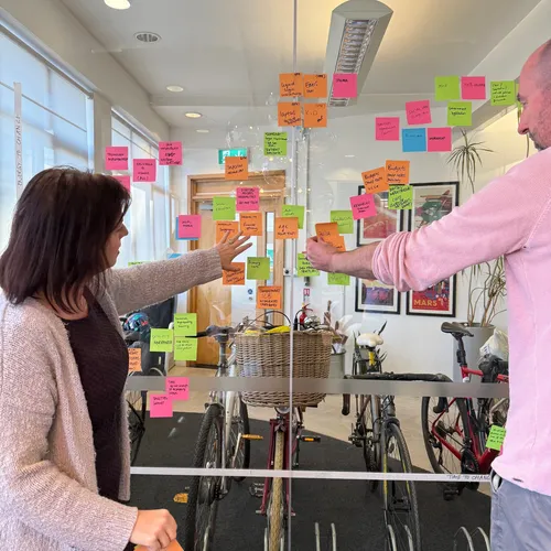 Two workshop participants stand at a glass wall adding colourful sticky notes to a growing cluster. Both are reaching and arranging ideas, reflecting an engaged, hands-on group activity.