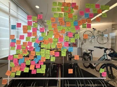 A glass wall covered in dozens of colourful sticky notes with handwritten ideas, seen inside an office with bikes and desks behind the glass.