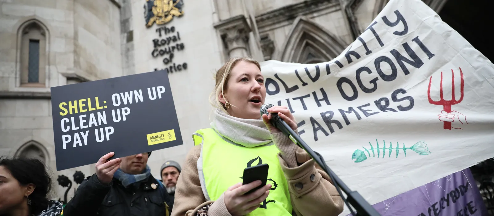 An Amnesty International campaigner wearing a fluorescent yellow vest speaks into a microphone outside the Royal Courts of Justice in London. Behind her, protesters hold a placard reading “Shell: Own Up, Clean Up, Pay Up” and a large banner reading “Solid