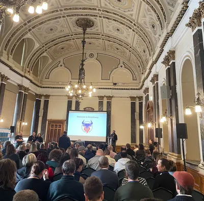 A large audience seated inside Birmingham City Council House, facing a stage with a screen reading “Welcome to BrumCamp” during UK Gov Camp.