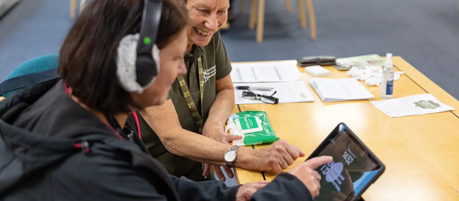 Two people sitting together at a table, smiling as they look at and interact with a tablet. One is wearing an RNID lanyard, and the other is wearing headphones.