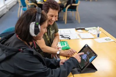 Two people sitting together at a table, smiling as they look at and interact with a tablet. One is wearing an RNID lanyard, and the other is wearing headphones.