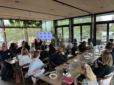 Lisa welcomes attendees. The venue is bright and modern, with large windows and greenery in the background.