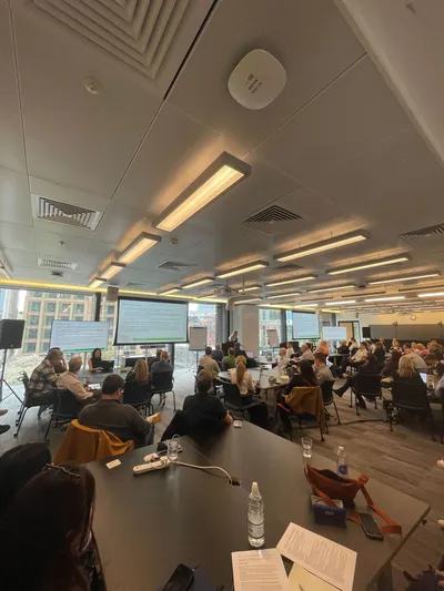 Attendees seated at round tables listening to a presentation during the LGRCamp workshop in a large conference room with presentation screens at the front.
