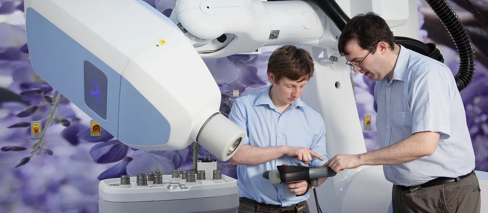 Two healthcare professionals demonstrate a piece of radiotherapy equipment, pointing to controls on a handheld device beside the large medical machine.