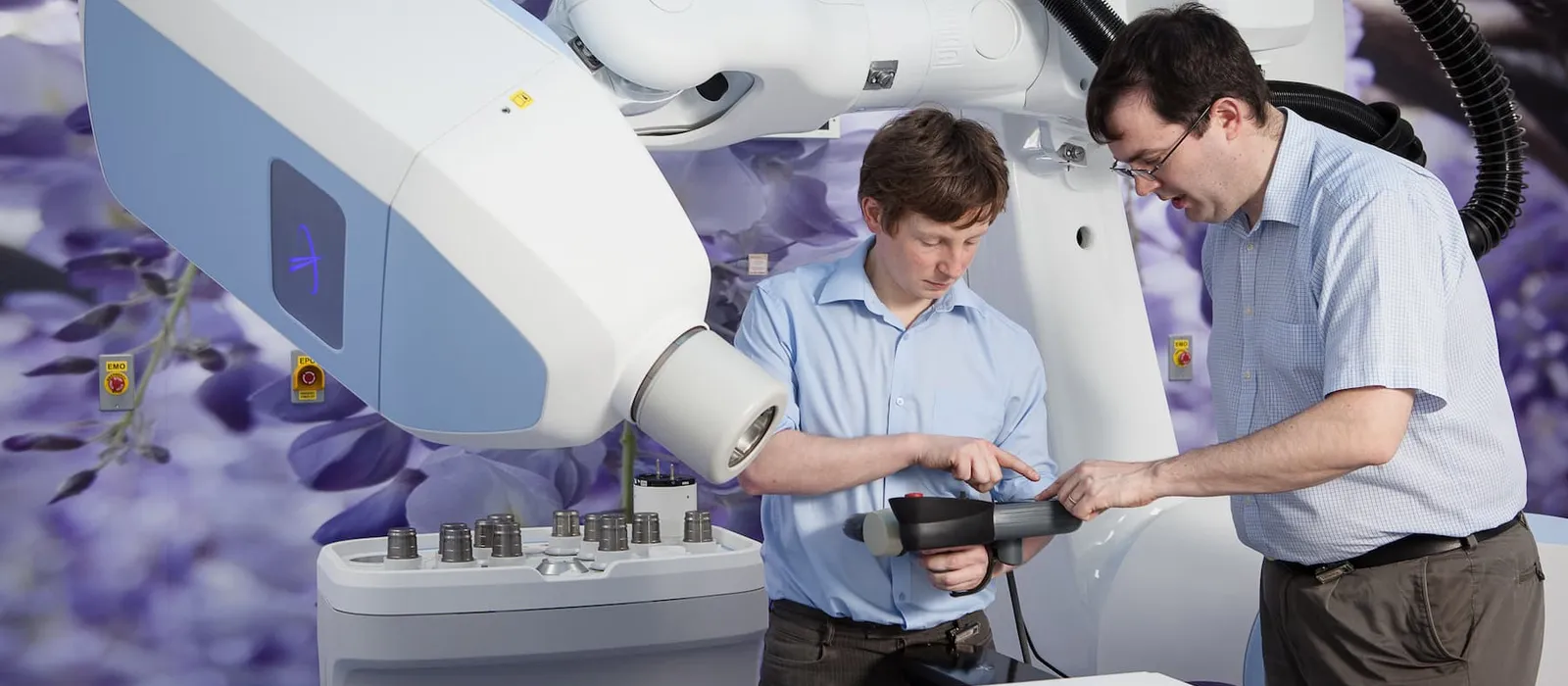 Two healthcare professionals demonstrate a piece of radiotherapy equipment, pointing to controls on a handheld device beside the large medical machine.