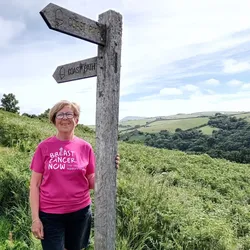 A person wearing a bright pink T-shirt stands beside a wooden signpost on a countryside hill, with a walking trail and rolling green landscape stretching into the distance.