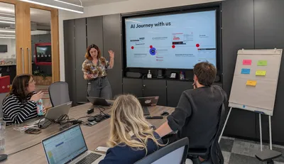 Iona stands at the front of a meeting room explaining a slide titled "AI Journey with us" on a large screen, while several colleagues seated around a conference table listen with laptops open. A flip chart with colourful sticky notes stands beside the screen.