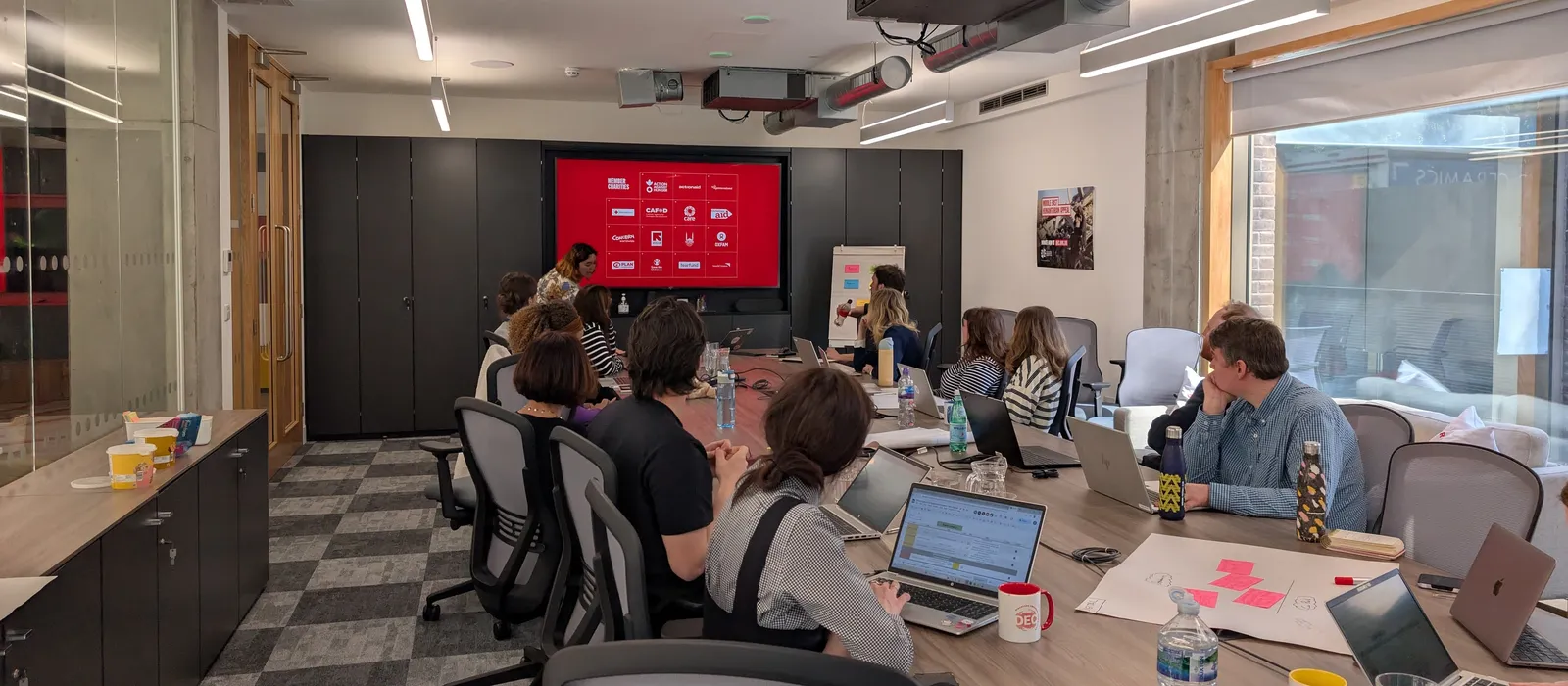 A group of people sit around a large conference table in a modern meeting room, listening to a presenter standing beside a large screen displaying a presentation. Several attendees have open laptops, notebooks and water bottles on the table.
