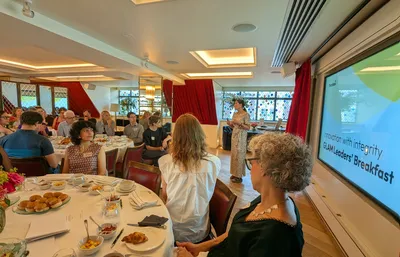 Lisa welcomes guests to Torchbox's GLAM Leaders' Breakfast, standing at the front of a light-filled room beside a large screen displaying the event title. Attendees are seated around breakfast tables with pastries, fruit and coffee as they listen to the opening remarks.