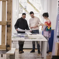 Three people stand around a large table in a bright studio space. They appear to be discussing the designs and materials on the table in front of them, smiling and engaged.