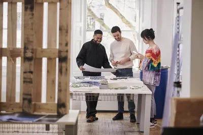 Three people stand around a large table in a bright studio space. They appear to be discussing the designs and materials on the table in front of them, smiling and engaged.