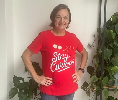 Dr Cath Biddle, stands smiling in front of a softly lit wall with plants, wearing a bright red top with the words “Stay Curious” in white lettering.