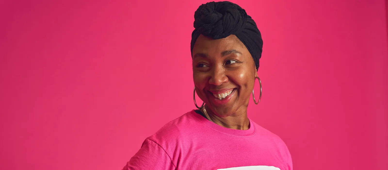 A smiling person wearing a bright pink Breast Cancer Now t-shirt and black headwrap stands against a pink background, looking to one side.
